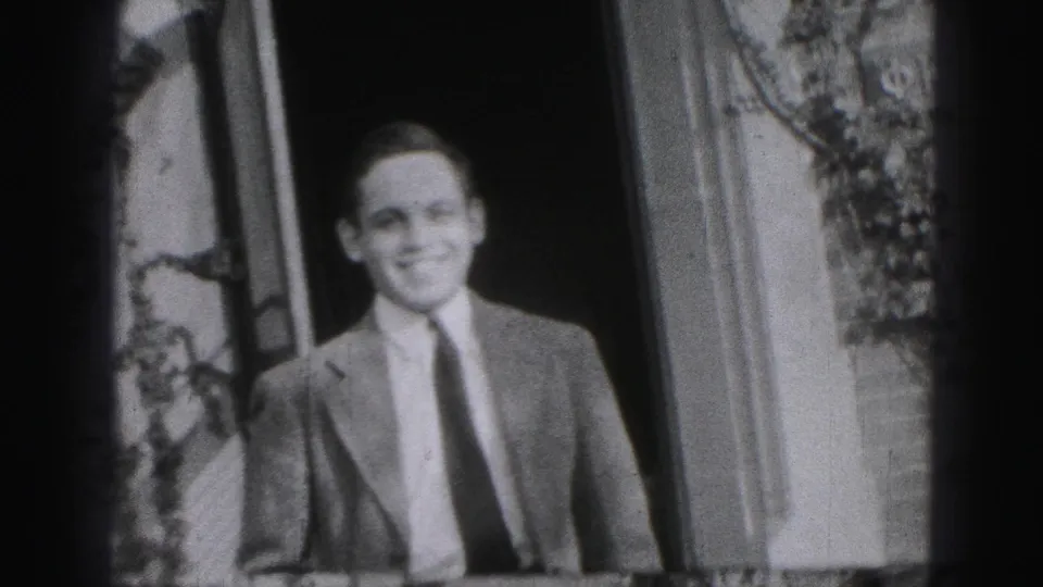 Smiling adult man in a suit and tie faces the camera outside a Harrisburg house in 1946 home-movie footage.