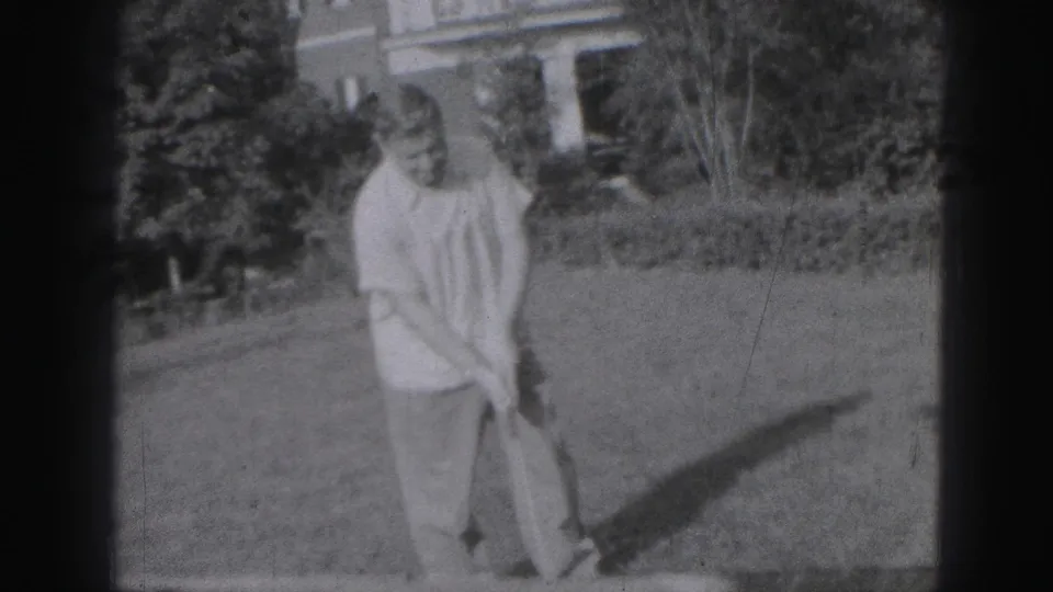 In 1946 Harrisburg home-movie footage, a man in a light shirt stands on a grassy yard with a golf club, trees and a house behind him.