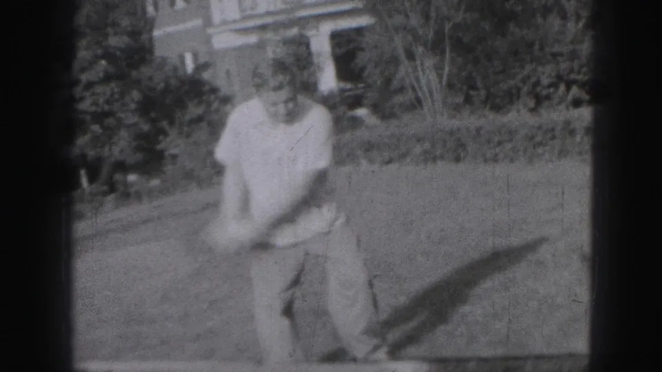 The golfer moves through his swing on a Harrisburg lawn, with motion blur, shrubs, and part of a house visible in the 8mm frame.