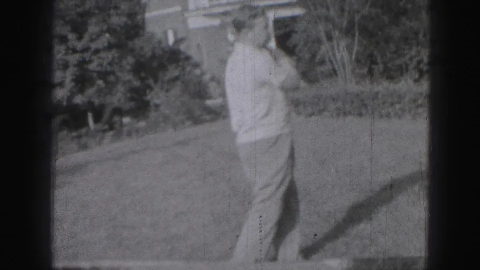 At the end of the shot, the man faces away after completing the golf swing, with the lawn, garden planting, and house exterior behind him.
