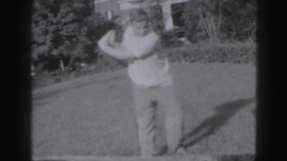 In Harrisburg 1946 home-movie footage, a man in light clothing stands on a lawn with a golf club near shrubs and a house.