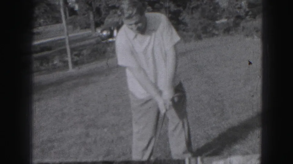 A man in light shirt and trousers bends over a golf club on a Harrisburg lawn, with trees and a house visible behind him.