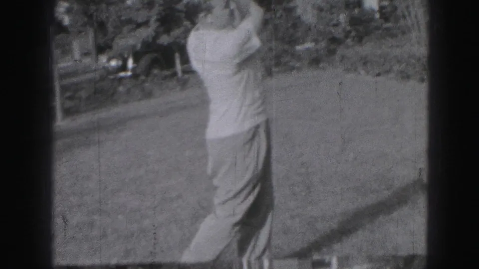 A man in light clothing follows through with a golf club on a grassy Harrisburg lawn, framed by shrubs and dark film edges.