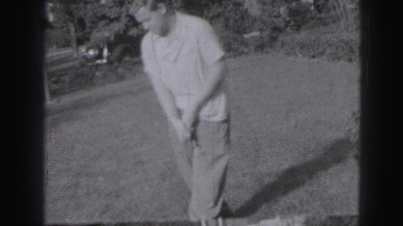 The golfer addresses the ball or ground with his club on grass, with shrubs and a parked vehicle visible in the background.