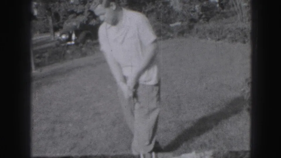 The golfer addresses the ball or ground with his club on grass, with shrubs and a parked vehicle visible in the background.