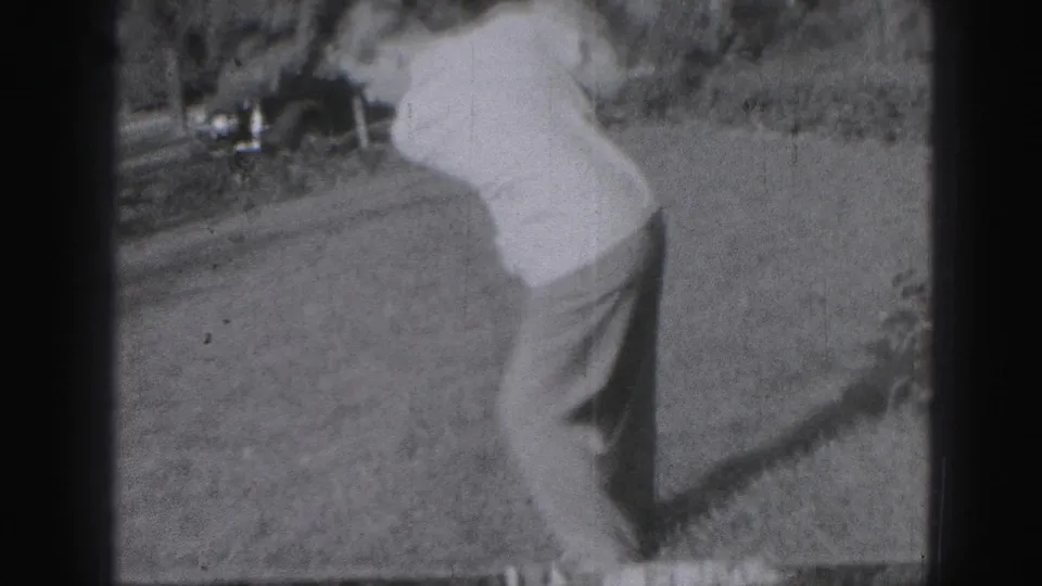 The golfer turns through his swing in black-and-white 8mm footage, with the grassy ground and background landscaping partly visible.