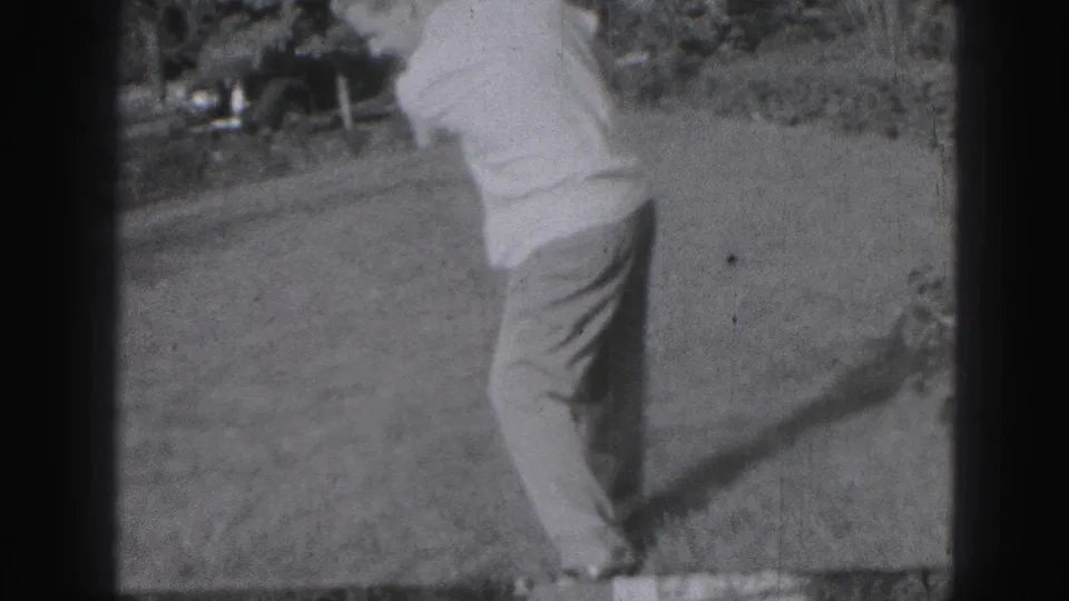 The golfer finishes his swing with his body turned away, framed by black-and-white 8mm grain, grass, and background shrubbery.
