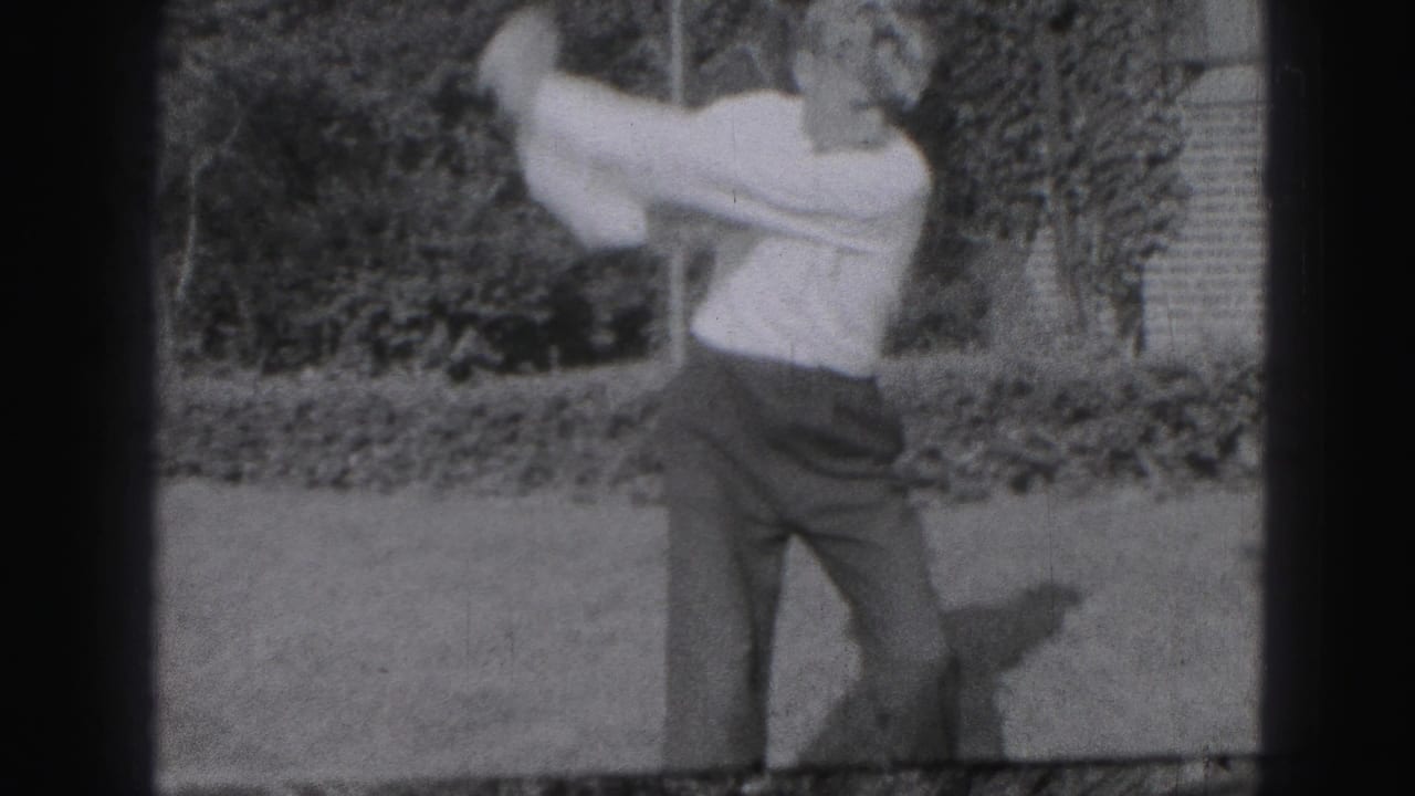 Man mid-swing with a golf club on a lawn near shrubs.