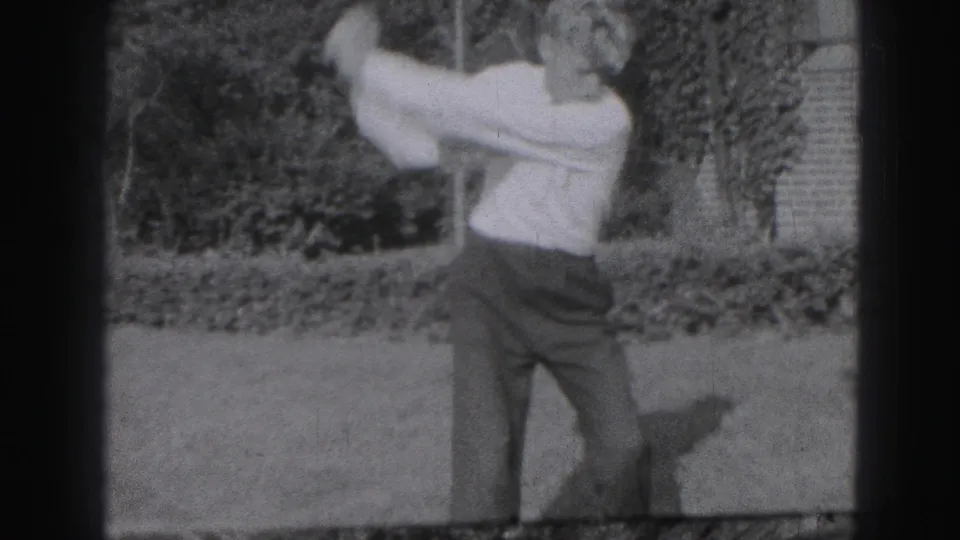 The man practices a golf swing on a Harrisburg lawn, with shrubs and a building edge behind him.