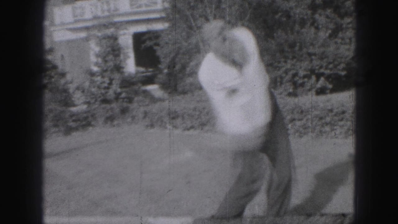 Man follows through on a golf swing on a lawn in front of shrubs and a house.