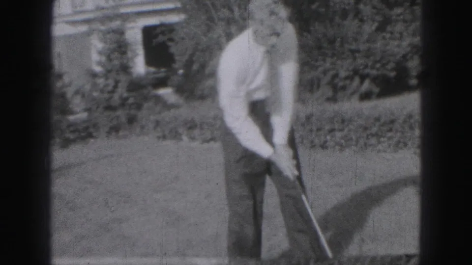 A black-and-white 8mm frame shows the man resetting or beginning another golf swing on the Harrisburg lawn.