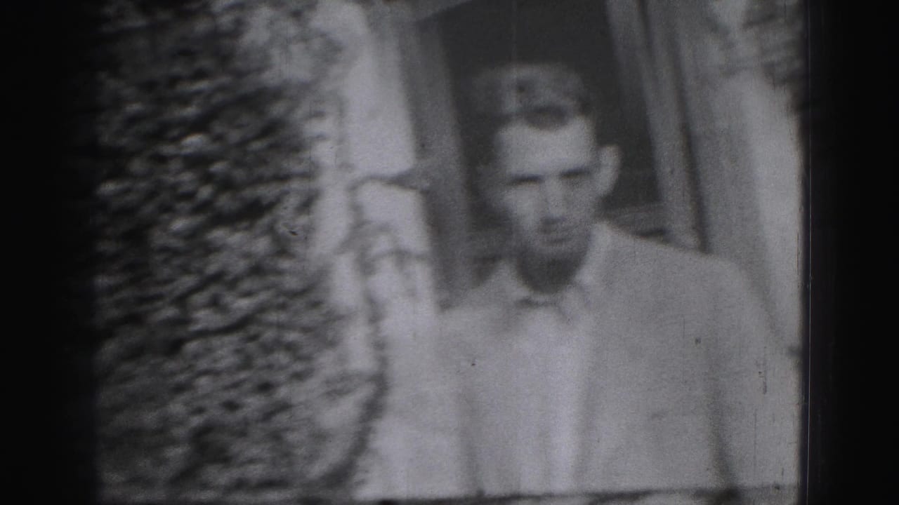 Young man in a light shirt walking near foliage and a wall.