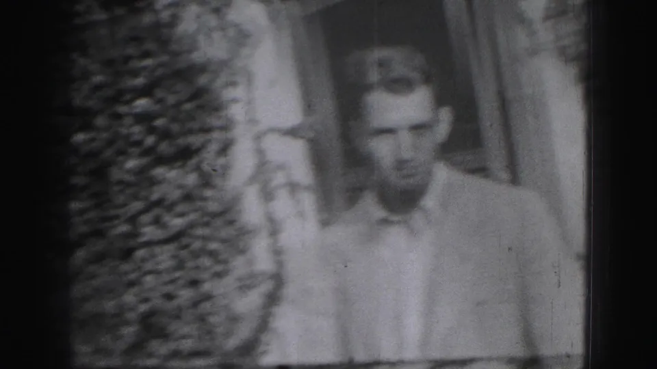 A young man in a light collared shirt appears beside leafy foliage and an exterior wall in the Harrisburg home-movie clip.