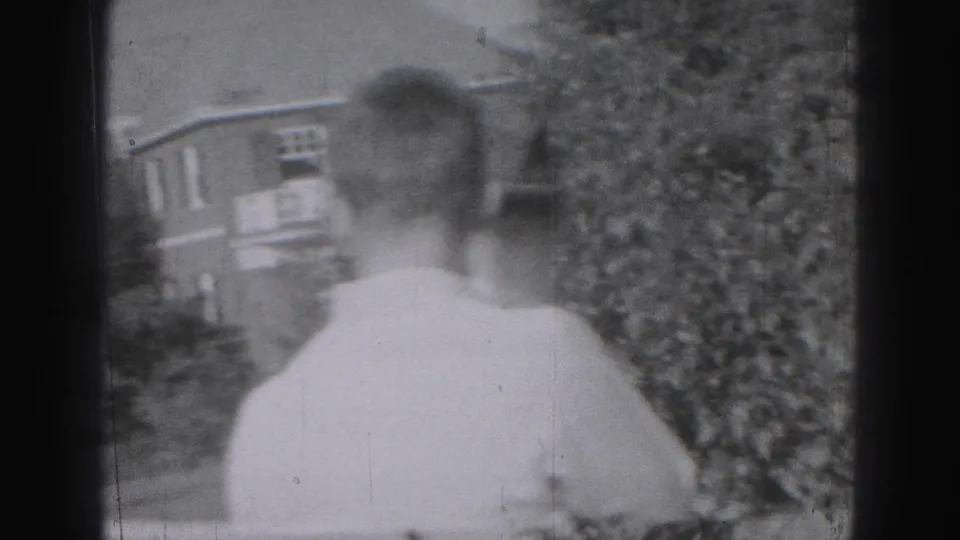 The camera catches the back of a young man in a light shirt, with shrubs and nearby residential buildings in view.