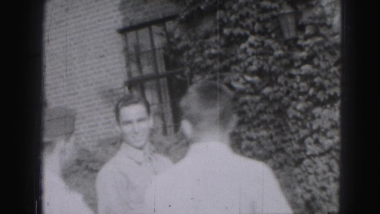 Two adult men near a brick wall and leafy garden, one facing the camera and one turned away.