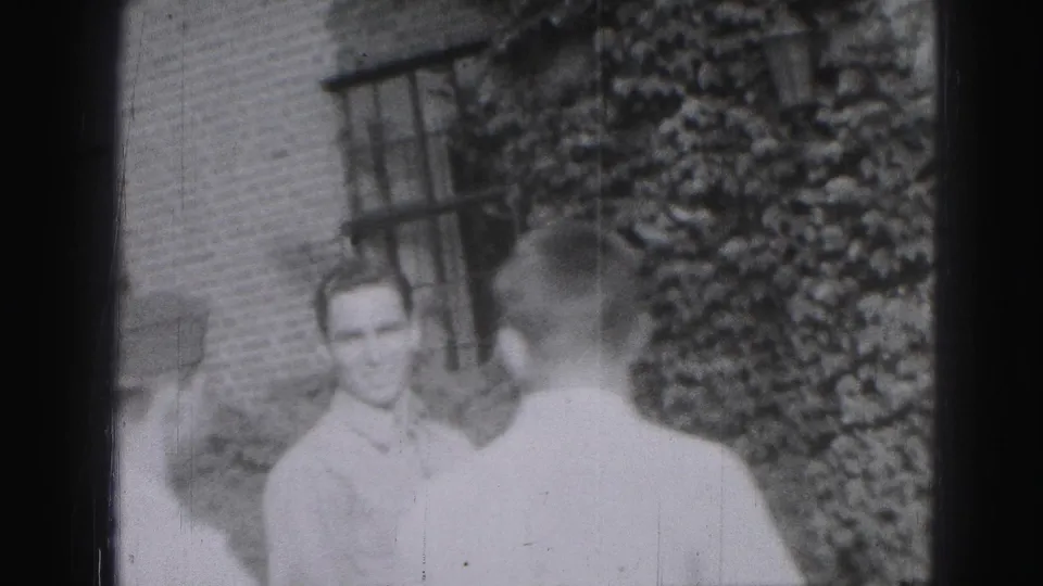 1946 Harrisburg home-movie frame showing two adult men near a brick building and ivy-covered garden area.