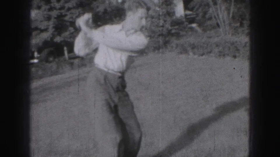 A man completes a full golf swing in a Harrisburg garden, with a lawn, shrubs, and 8mm film grain visible.