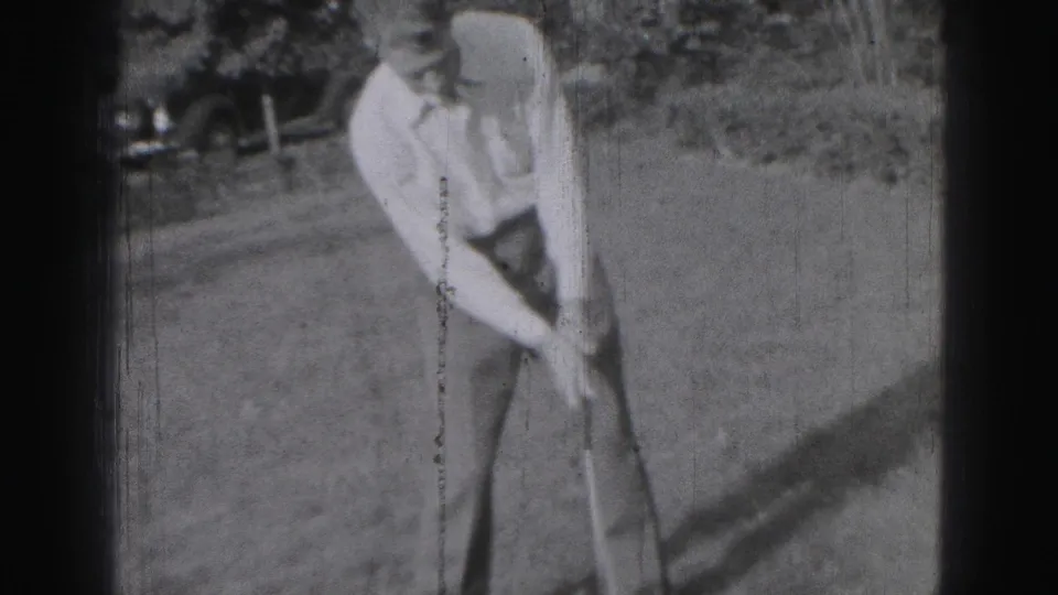 Black-and-white 8mm frame from Harrisburg in 1946 showing a golfer addressing the ball, with an older car visible behind the lawn.