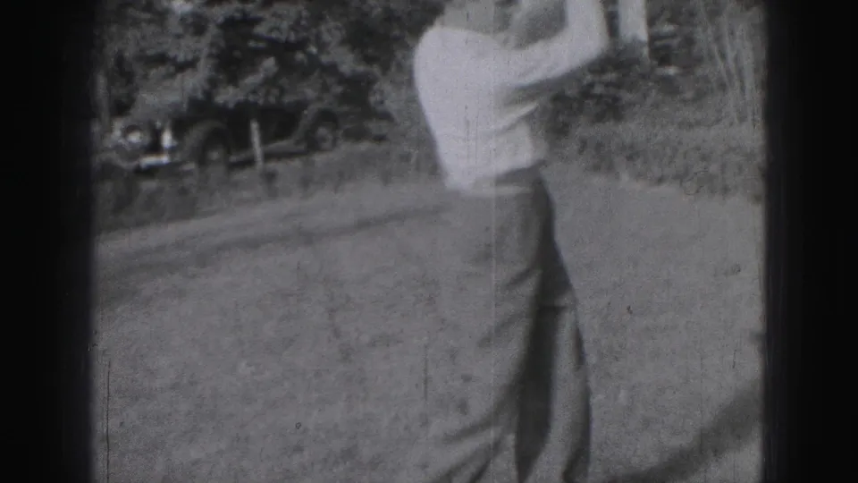 Cropped 1946 Harrisburg home-movie frame showing the golfer's lower body on the lawn during a golf swing, with trees and a car behind.