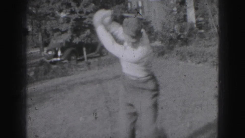 Soft black-and-white 8mm frame from 1946 Harrisburg showing a golfer finishing a swing, with a parked older car in the background.