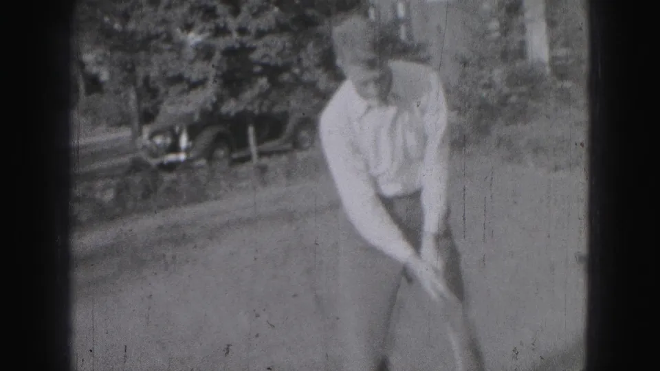 Adult golfer in light clothing bends into his swing on a Harrisburg grass course, with a 1940s car in the background.