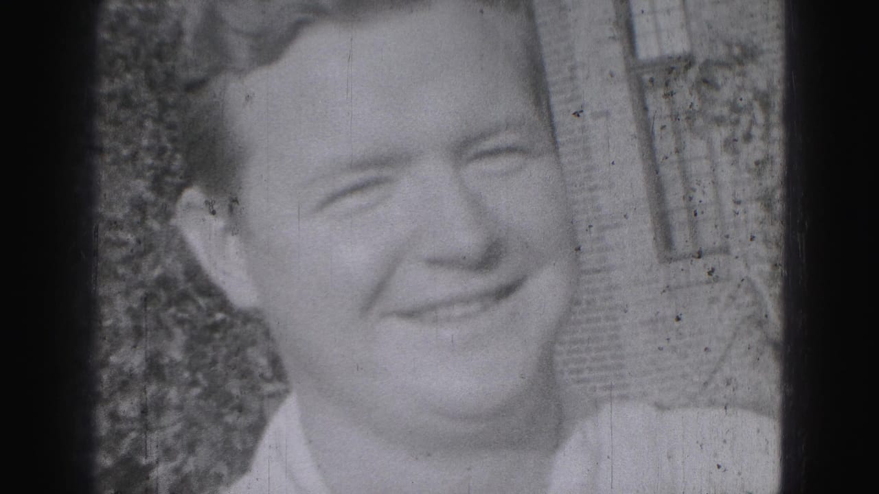 Close-up of a smiling young man outdoors beside shrubs and brickwork.