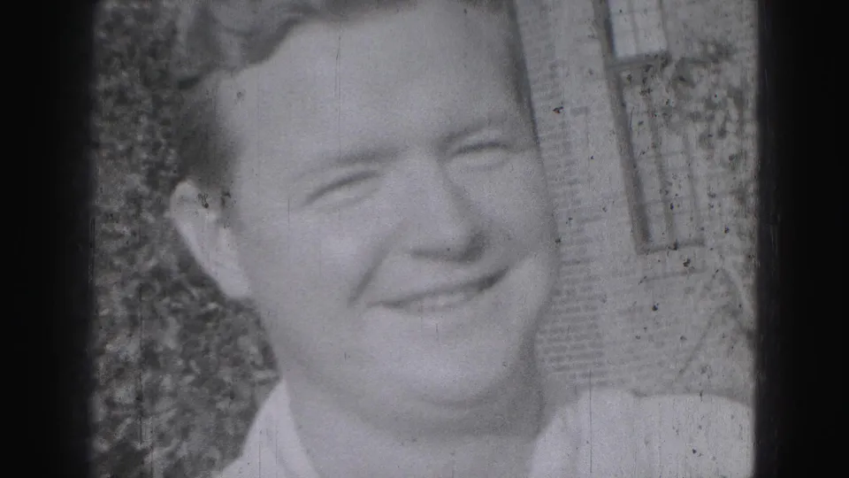 Close-up of a smiling young man outdoors in the Harrisburg home-movie footage, with shrubs and a brick building behind him.