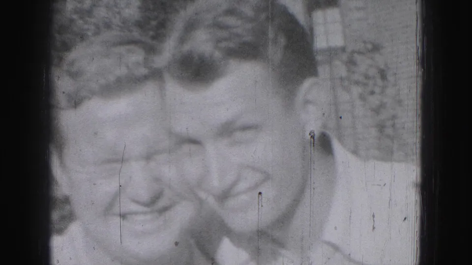Two young men smile close together for the camera in the 1946 Harrisburg home movie, framed by shrubs and a brick building.