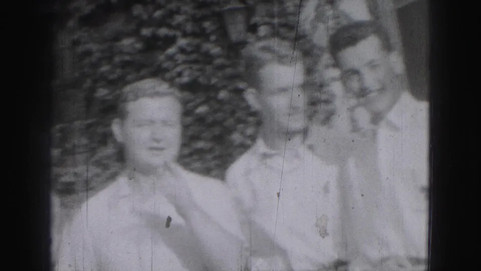 In the 1946 Harrisburg home-movie frame, three young men stand outdoors while one touches his collar or neckline.