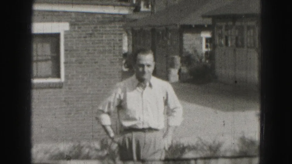 1946 Harrisburg 8mm frame showing an adult man in a light shirt and trousers standing outside a brick house.