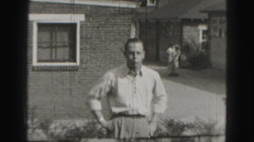 In the Harrisburg 1946 home-movie frame, a man stands on a lawn before a brick house with another adult and small dog behind him.
