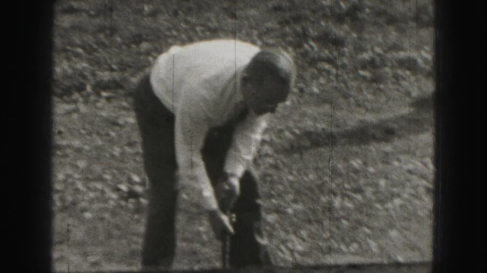 A man in a light shirt and dark trousers bends over on a gravelly outdoor area in the 1946 Harrisburg home movie.