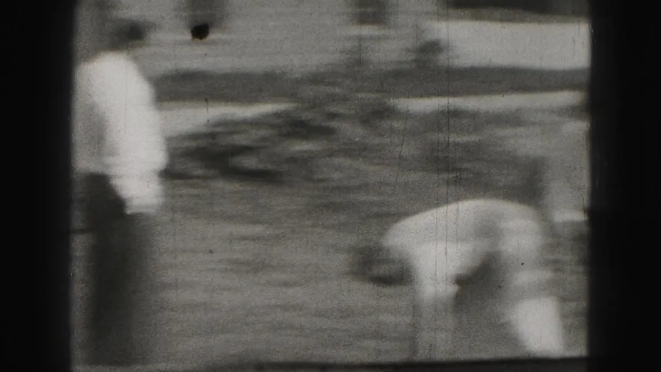 Soft black-and-white frame from the Harrisburg clip with one man bent over the yard during croquet and another figure nearby.