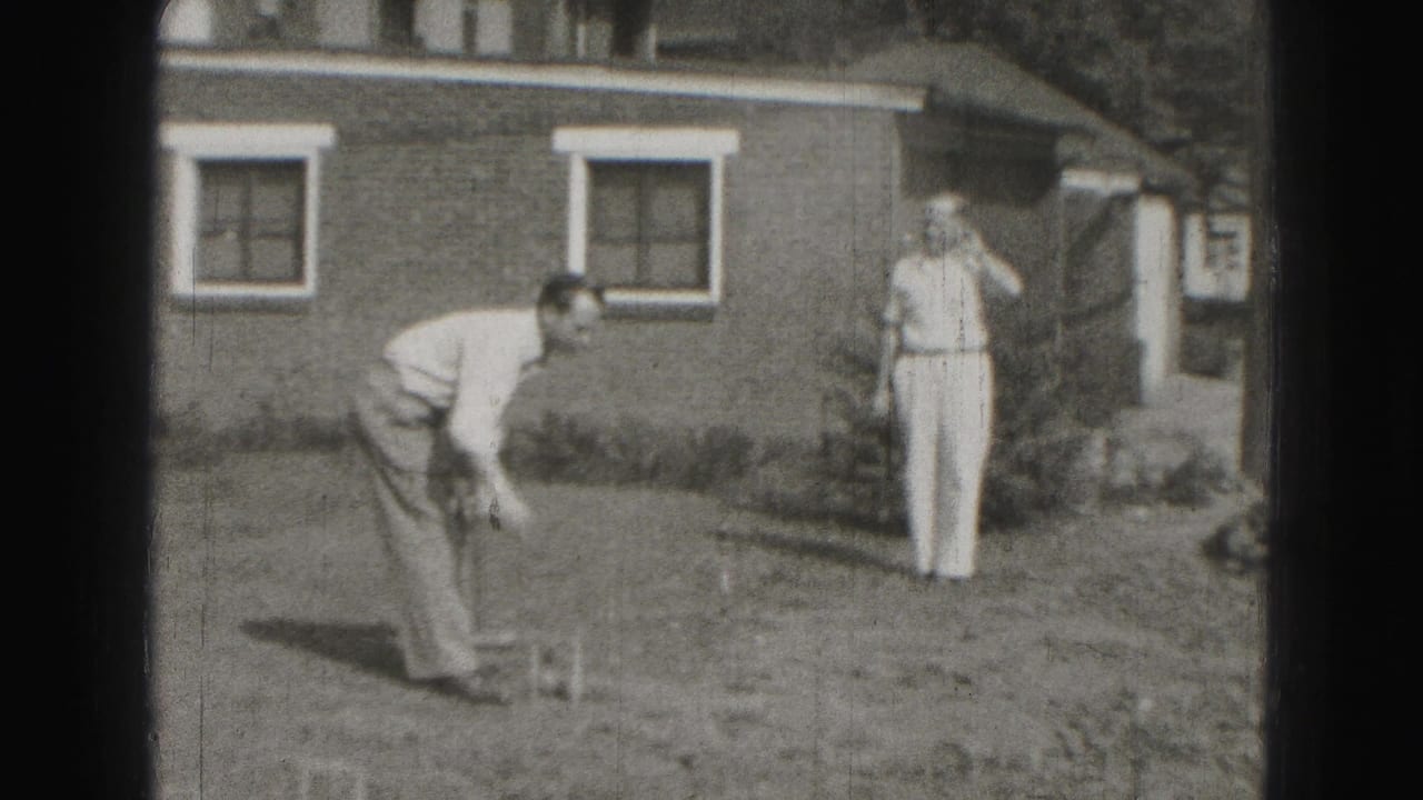 Two men on a backyard lawn with a croquet mallet and a brick house behind them.