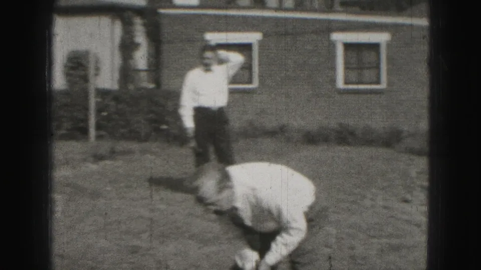 In 1946 Harrisburg home-movie footage, one adult man bends toward a croquet shot as another stands near a house and lawn.