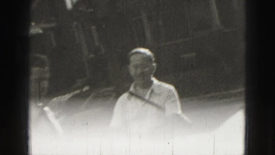 Adult man in light clothing stands outdoors in the Harrisburg home-movie croquet scene, with yard and background structures behind him.