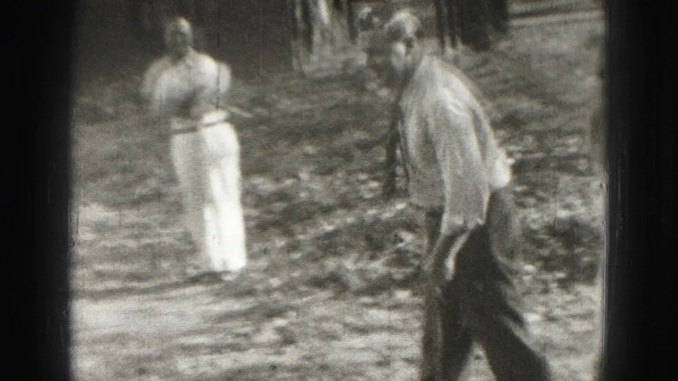 Two adult men in light shirts and trousers appear in a 1946 Harrisburg yard during a croquet game, one moving past the camera area.