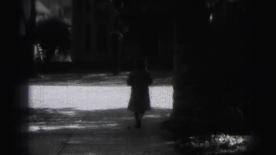 Black-and-white 8mm frame from the 1946 Harrisburg clip showing a distant pedestrian on a shaded sidewalk near trees and a building entrance.