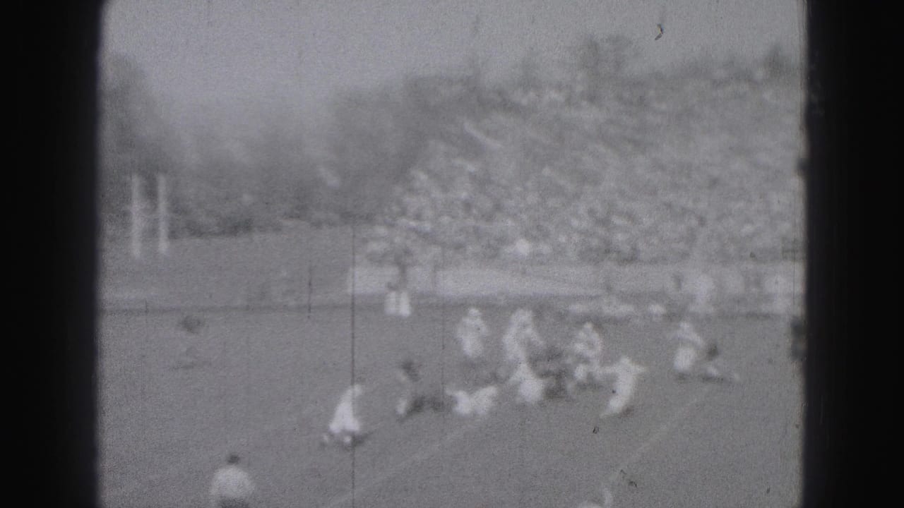 Players in light uniforms move and fall during football action near the center of the field.