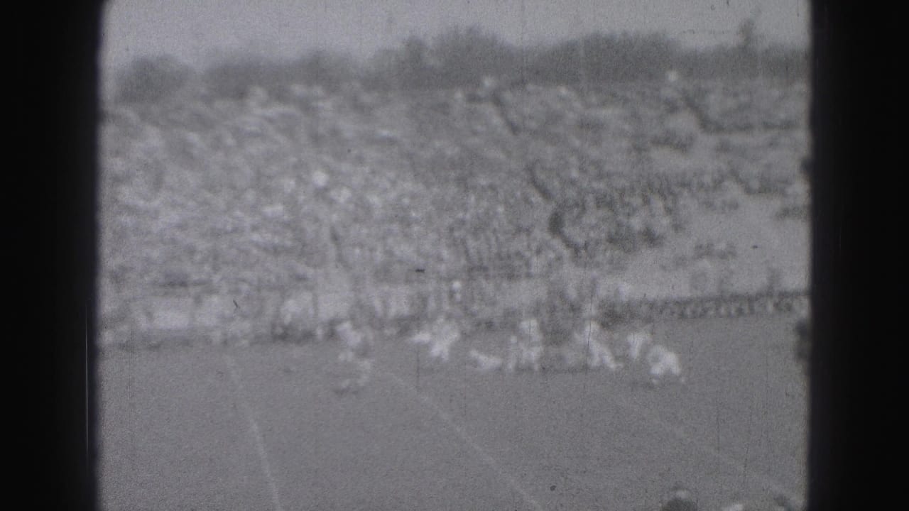 Soft black-and-white view of players near the sideline with a large crowd in the stands.