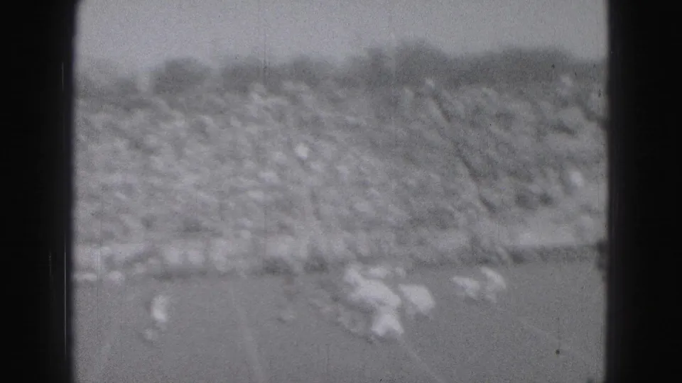 Black-and-white 8mm frame from the Harrisburg game, showing blurred football players below a packed stadium crowd.