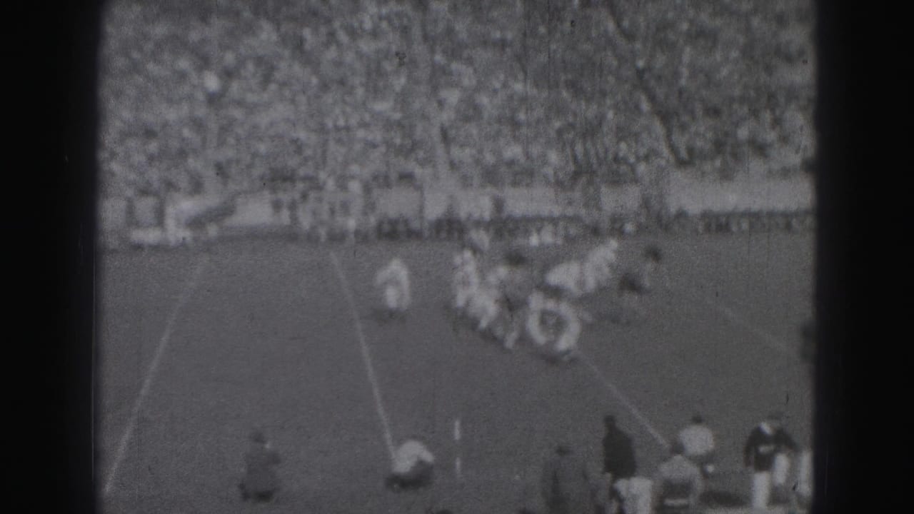 Football players collide on a grass field with spectators in bleachers behind them.