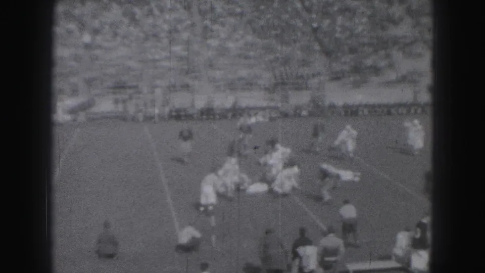 A 1946 Harrisburg high school football play continues as players gather around the ball with bleachers and spectators beyond the field.