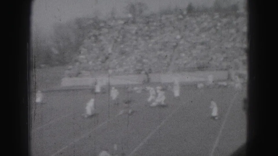 The football game continues in Harrisburg in 1946, with players moving across the marked field and a large spectator crowd behind them.