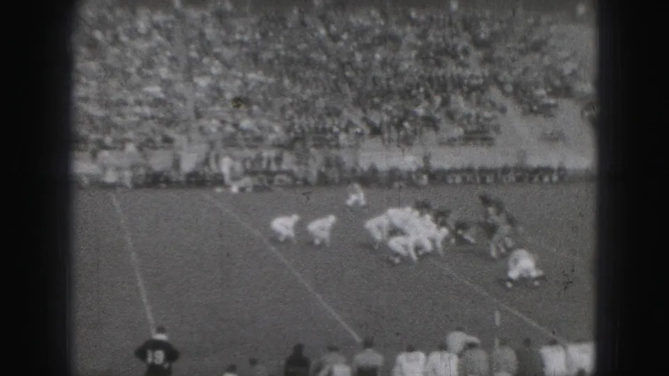 Players line up for a football snap in 1946 Harrisburg, with dark and light uniforms, yard lines, sideline personnel, and a large crowd beyond.