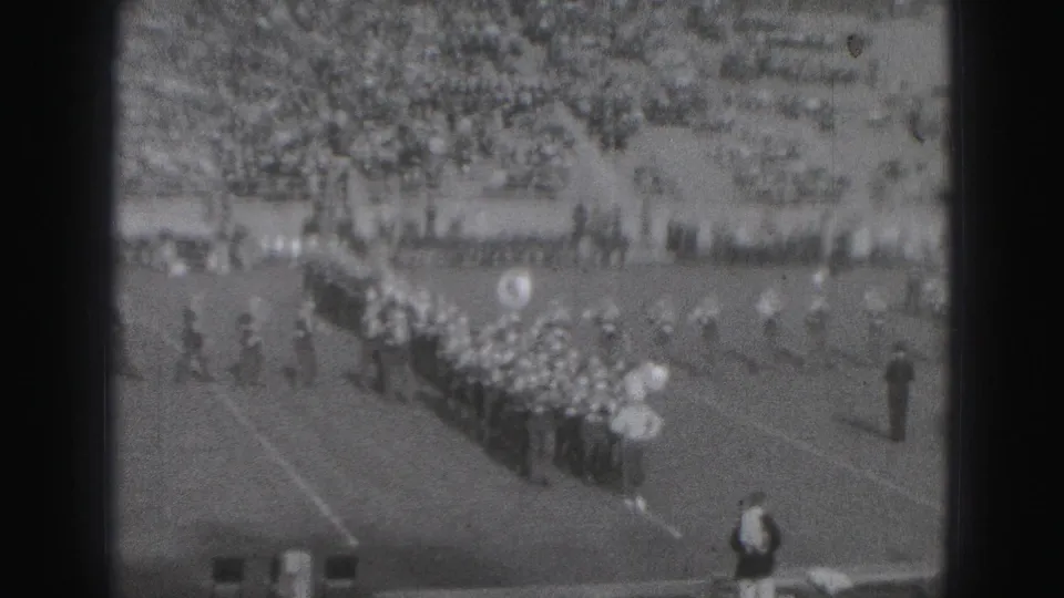 Marching band members move in formation across a Harrisburg football field while spectators watch from the stands.