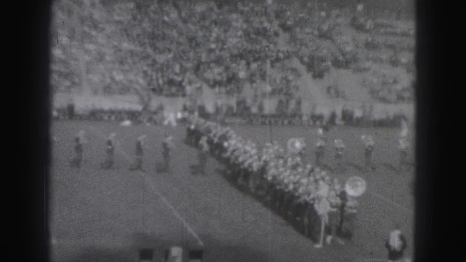A distant 8mm frame shows the marching band in diagonal formation on a Harrisburg football field with spectators in the background.