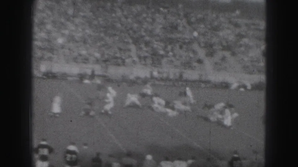 Home-movie frame of 1946 Harrisburg rugby play, showing players spread across the field in front of crowded spectator stands.