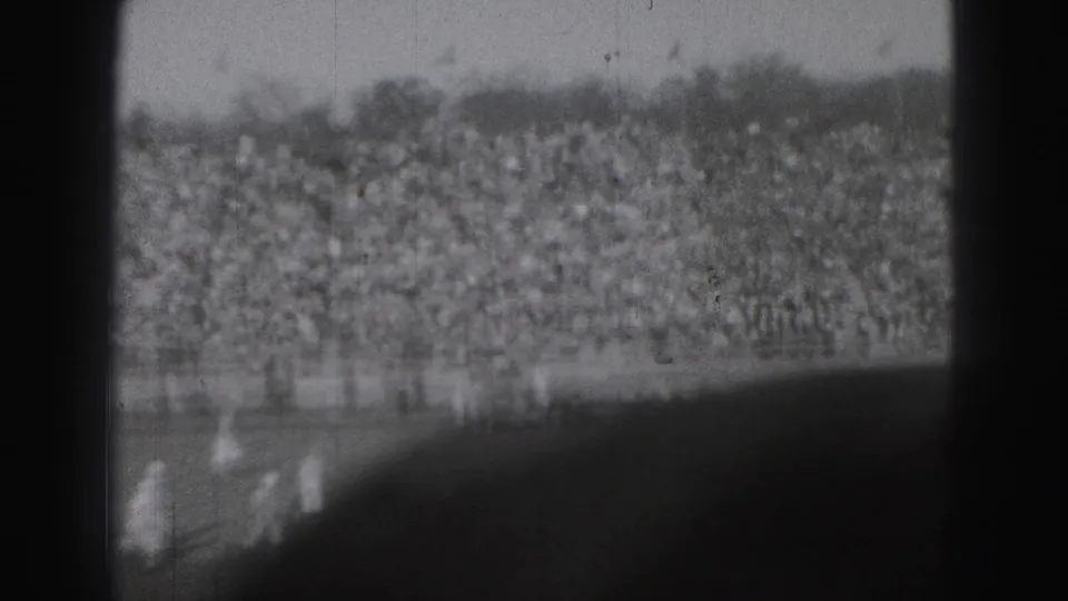 The camera frames the crowded Harrisburg stands, with blurred sideline figures and field edge visible in front.