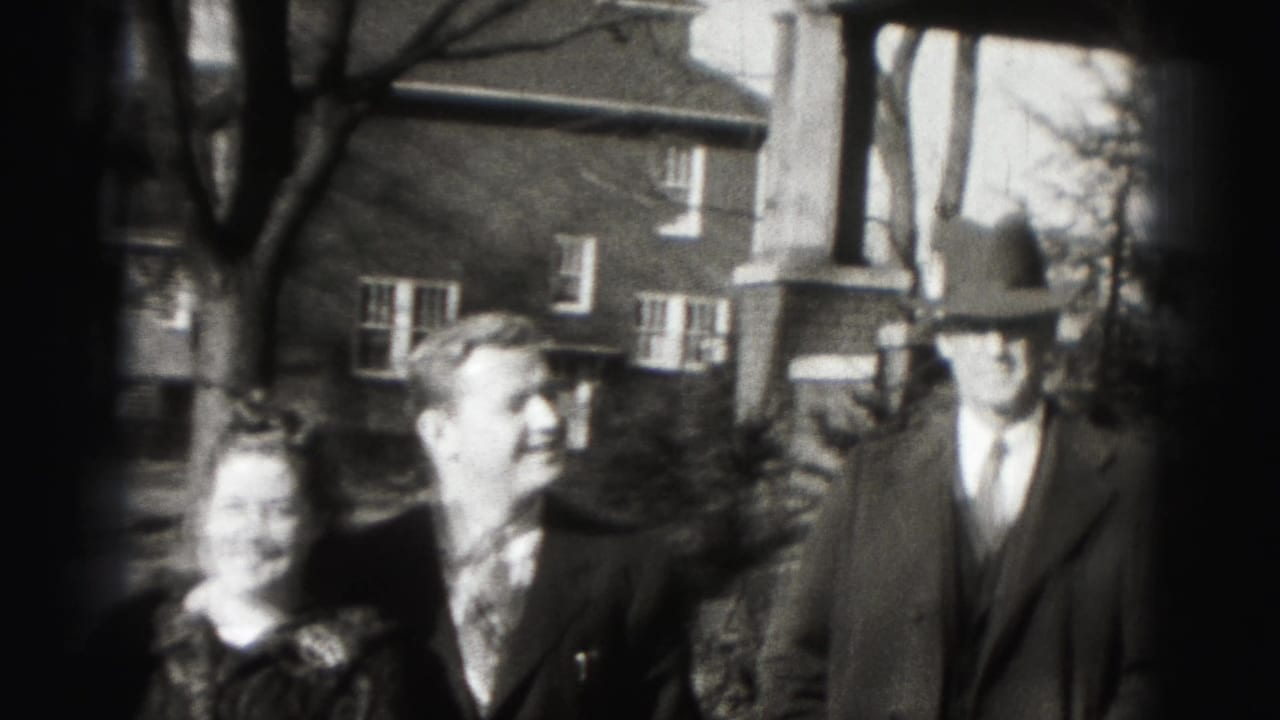 Three adults in formal clothing stand outside a brick house near a porch.